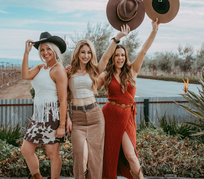 Three smiling women in boho country outfits and cowgirl boots pose in front of vineyard rows and a corrugated metal fence, two holding wide-brim hats aloft against a blue sky.