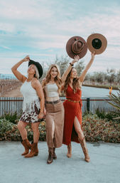 Three women in boho-cowgirl outfits and cowboy boots, smiling and raising hats beside vineyard rows under a blue sky