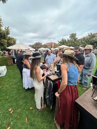 Crowded outdoor craft market on a cloudy day, shoppers browse a hat and accessories stall on green grass—people in summer dresses, denim vests and wide‑brim hats with umbrellas and vendor tents in the background.