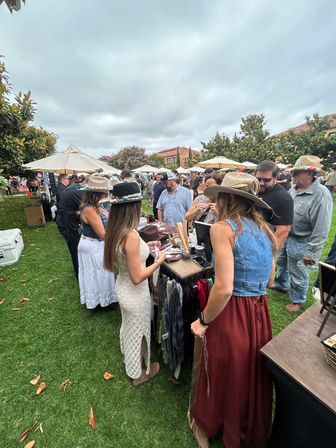 Crowded outdoor craft market on a cloudy day, shoppers browse a hat and accessories stall on green grass—people in summer dresses, denim vests and wide‑brim hats with umbrellas and vendor tents in the background.
