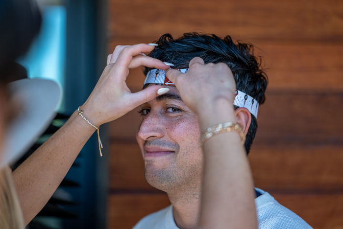 Close-up of a smiling man having his head circumference measured with a paper tape measure by hands wearing bracelets, wooden wall background — hat fitting/head sizing scene.