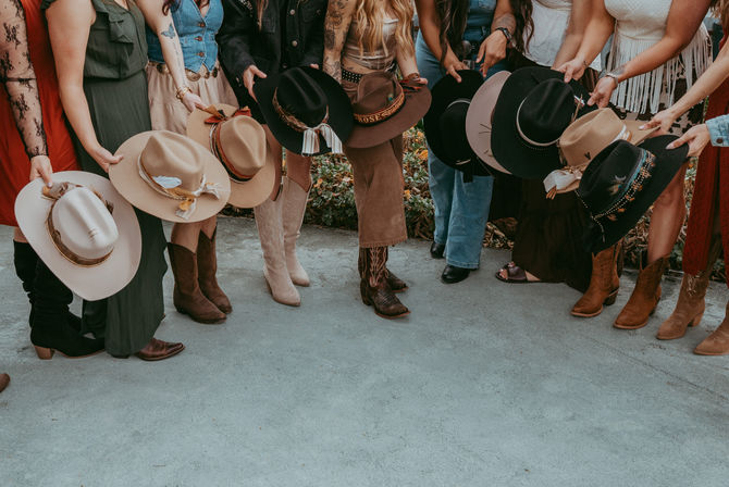 Row of people outdoors holding cowboy hats and wearing western boots in boho-western styles — denim, fringes and earth-tone hatbands