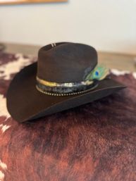 Dark brown felt cowboy hat with a wide brim, decorative leather band and peacock feather accent, resting on a brown-and-white cowhide rug — rustic Western vibe.