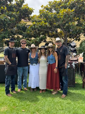 Group photo of six people in cowboy hats and western-style outfits standing on a grassy lawn in front of vendor tables and leafy trees at an outdoor craft market.
