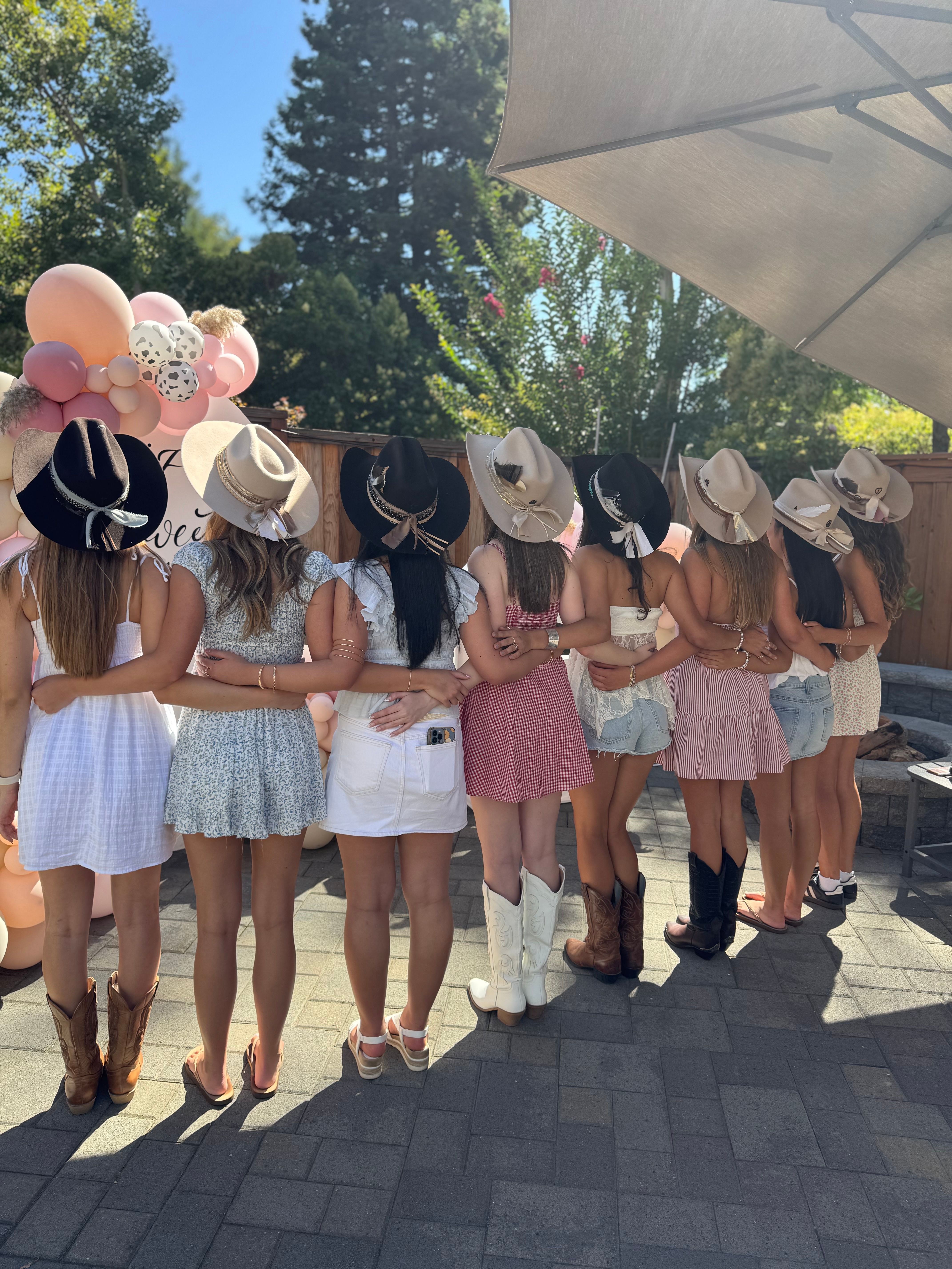 Row of friends on a sunny backyard patio with arms around each other, wearing cowboy hats and boots and casual summer dresses, posed in front of a pink balloon arch for an outdoor country-themed celebration