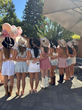 Row of friends on a sunny backyard patio with arms around each other, wearing cowboy hats and boots and casual summer dresses, posed in front of a pink balloon arch for an outdoor country-themed celebration