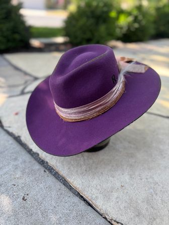 Purple wide-brim felt fedora with layered velvet ribbon and feather accent resting on a stone patio, stylish fashion accessory