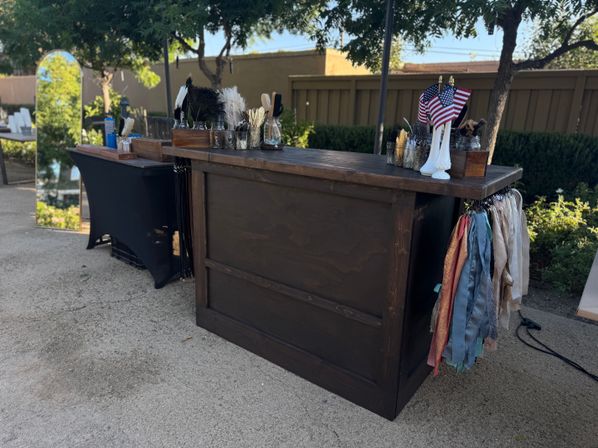 Rustic wooden outdoor bar and prop station with jars of feathers, colorful fabric ribbons hanging at the side, small American flags in a white vase, a tall mirror and trees along a backyard fence for an outdoor event.
