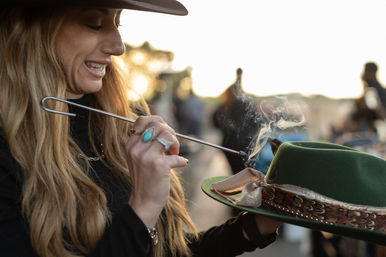 Smiling woman steaming and reshaping a green felt hat with feather trim outdoors at sunset, close-up of her hand with a turquoise ring and rising steam.