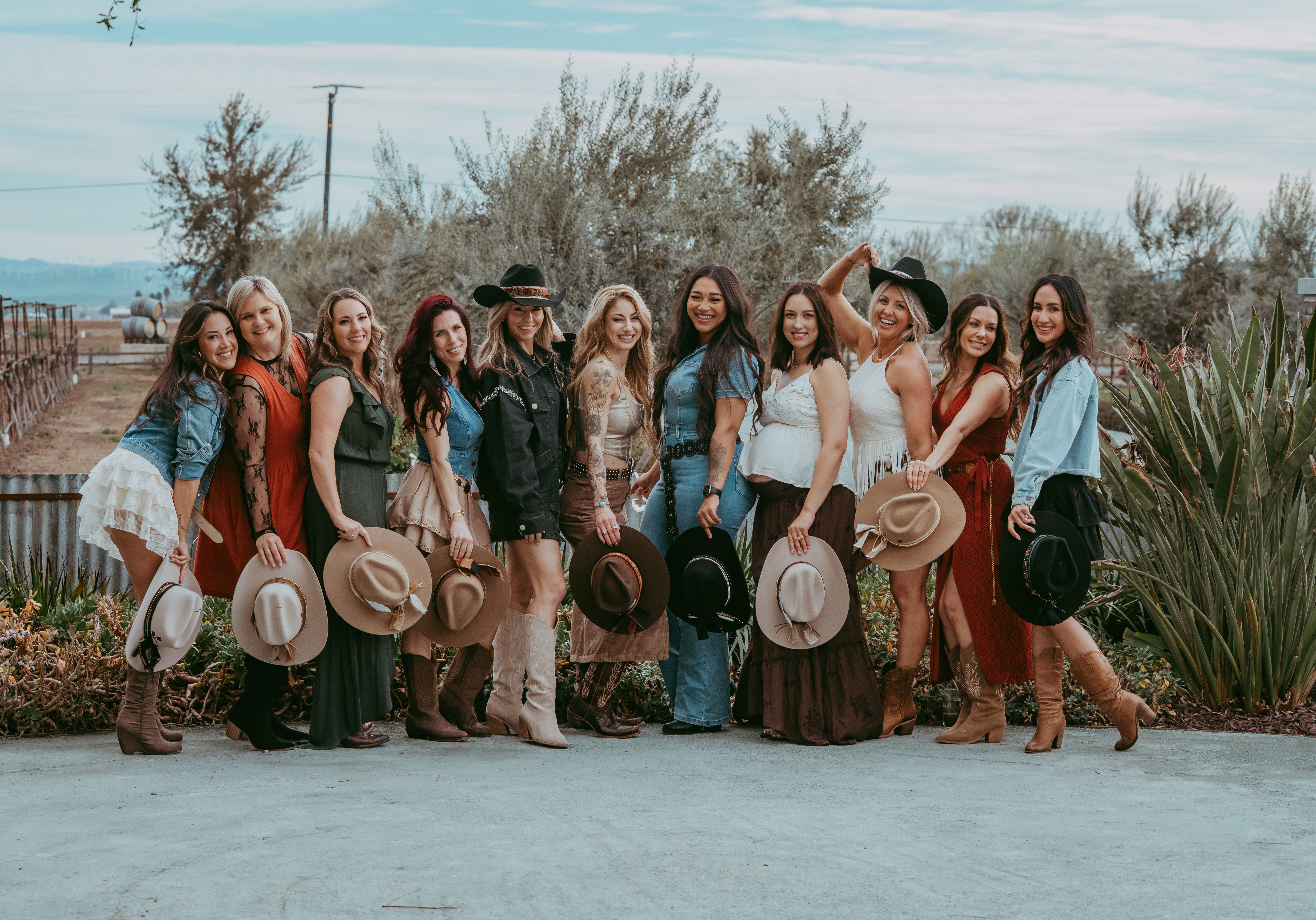 Twelve women in cowgirl outfits and boots pose outdoors on a rural ranch, each holding a wide-brim hat and smiling against trees, fencing, and open countryside.