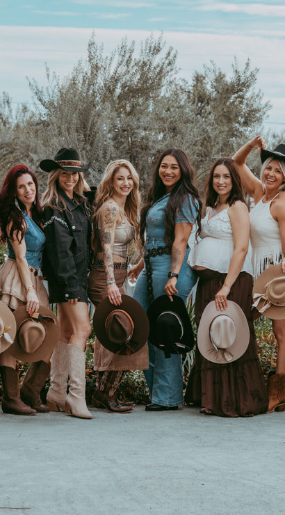 Twelve women in cowgirl outfits and boots pose outdoors on a rural ranch, each holding a wide-brim hat and smiling against trees, fencing, and open countryside.