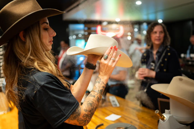 Person with tattooed arm steam-shaping a white cowboy hat at a western hat shop counter, neon bull sign and customers blurred in the background