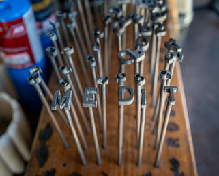 Close-up of metal letter stamps on long steel rods in a wooden rack spelling 'MEDIT', artisan metalworking tools on a workshop bench