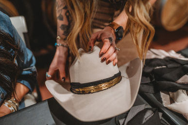 Hands with rings and long brown nails placing a cream cowboy hat with a black-and-gold band on a table in a rustic western boutique setting