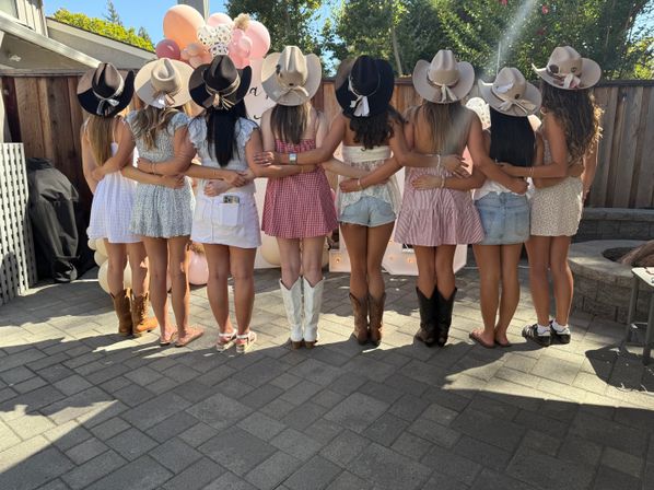 Back view of nine friends with arms around each other on a sunny backyard patio, wearing cowboy hats, summer dresses or shorts and cowgirl boots or sandals, posed in front of pink balloon decorations for a western‑themed outdoor celebration.