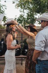 Outdoor backyard western-style reception: woman in white lace dress holding champagne has a white cowboy hat placed on her head by a friend in a brown hat, stacked cowboy hats and string lights in the background.