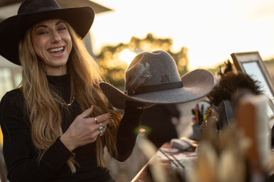 Smiling woman in a black hat holding a gray felt cowboy hat with feather trim and braided band at an outdoor stall during golden hour