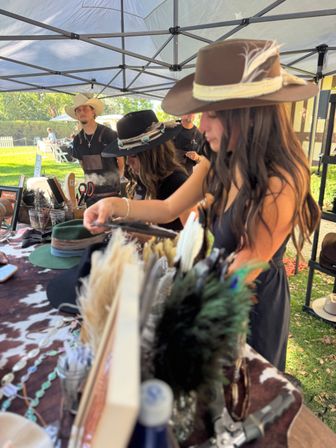 Outdoor craft market hat booth in a sunny park — two people trying on handcrafted cowboy hats with feather and turquoise bands under a canopy, table display of hat trims, feathers and accessories.