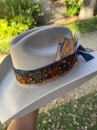 Wide-brimmed white felt fedora with a blue ribbon, layered pheasant feathers and gold chain trim, a Jack of Spades pin-up playing card tucked in the band, photographed outdoors over a grassy backyard.