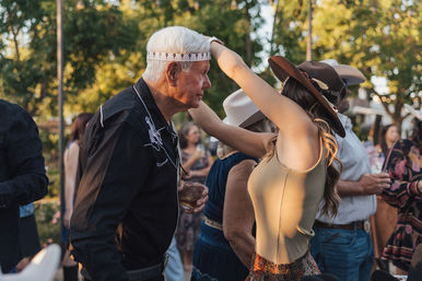 Woman in a brown hat playfully places a patterned headband on a gray-haired man at an outdoor country-style gathering, surrounded by guests in Western hats and warm evening light.