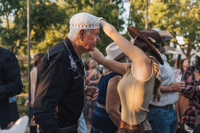 Woman in a brown hat playfully places a patterned headband on a gray-haired man at an outdoor country-style gathering, surrounded by guests in Western hats and warm evening light.