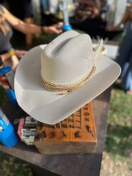 Cream felt cowboy hat with braided gold and leather hatbands and a feather accent, resting on a wooden display block at an outdoor market
