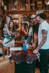 Four friends laughing in a rustic western shop trying on cowboy hats at a cowhide-covered counter with barrels and vintage signs in the background.