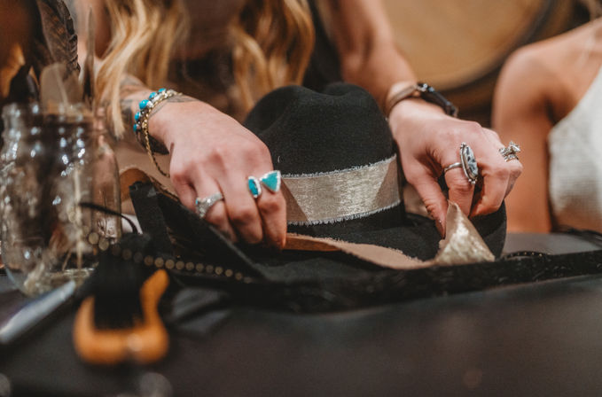 Close-up of hands wearing turquoise rings and bracelets adjusting a black fedora with a silver band on a worktable, boho accessories and glass jars nearby