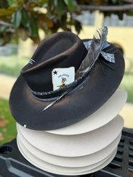 Black felt fedora with a Joker playing card and long gray feather tucked into a patterned hatband, displayed on stacked beige hat blocks outdoors with blurred greenery in the background.