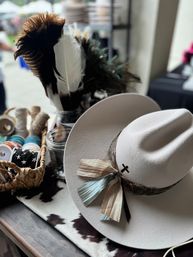 Stylish cream felt cowboy hat with feather-and-ribbon band on a cowhide-patterned table beside jars of assorted feathers, ribbons, and trim at an outdoor artisan market
