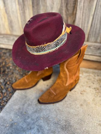 Burgundy felt wide-brim western hat with a feather-and-bead hatband resting on tan suede cowboy boots against a rustic wooden fence