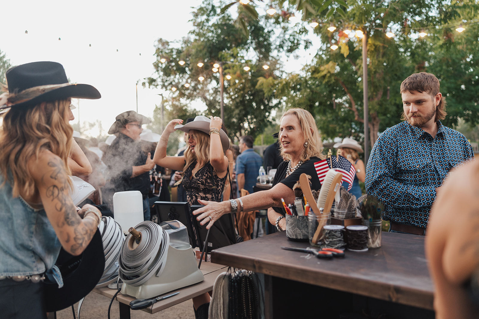 Lively outdoor country festival at dusk with people trying on cowboy hats at a vendor booth, grooming tools and small American flags on the table under string lights