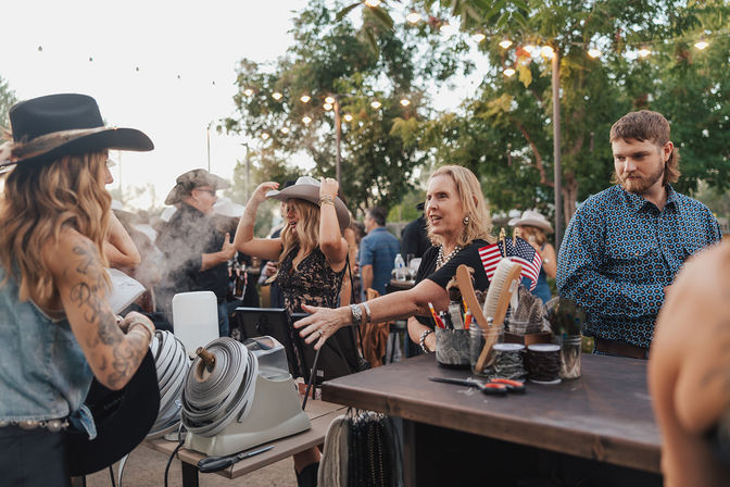 Lively outdoor country festival at dusk with people trying on cowboy hats at a vendor booth, grooming tools and small American flags on the table under string lights