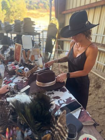 Woman in a black dress and wide-brimmed cowboy hat decorating a brown felt cowboy hat with a gold band on a cowhide-covered work table inside a rustic barn, surrounded by hatmaking tools, ribbons and stacked hats.