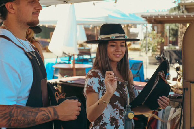 Smiling woman wearing a turquoise-banded wide-brim hat examines another hat at a sunny outdoor artisan market stall while a tattooed vendor in an apron assists.