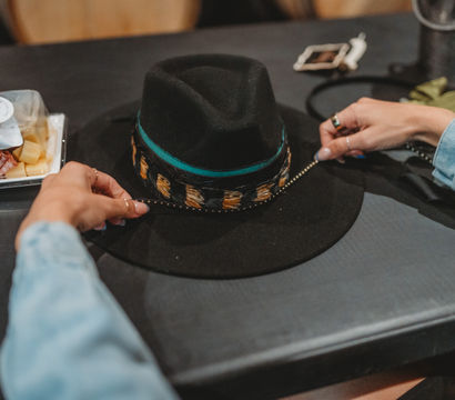 Hands with rings adjust a stylish black felt fedora with a turquoise and leopard-print band on a dark table beside a snack tray and smartphone