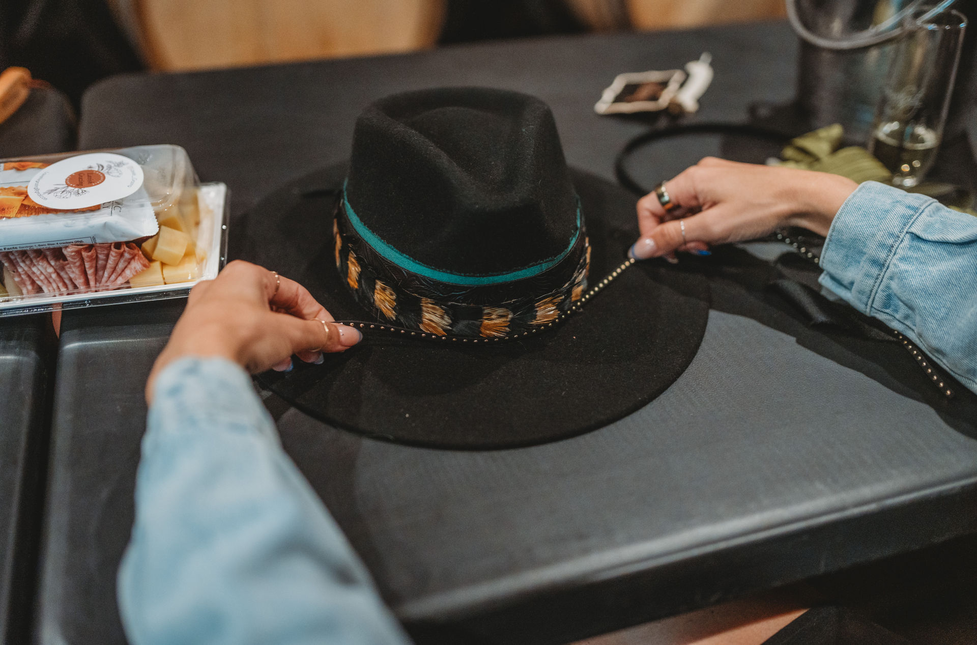 Hands with rings adjust a stylish black felt fedora with a turquoise and leopard-print band on a dark table beside a snack tray and smartphone
