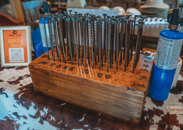 Wooden block holding dozens of metal stamping punches upright on a cowhide-covered table, with a blue butane torch nearby — leathercraft stamping tool display.