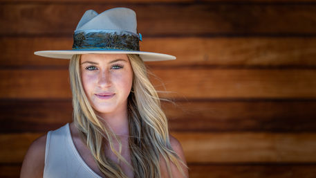 Sunlit portrait of a blonde woman with long hair and freckles wearing a wide-brimmed hat with a feather band, posed against warm wooden planks.