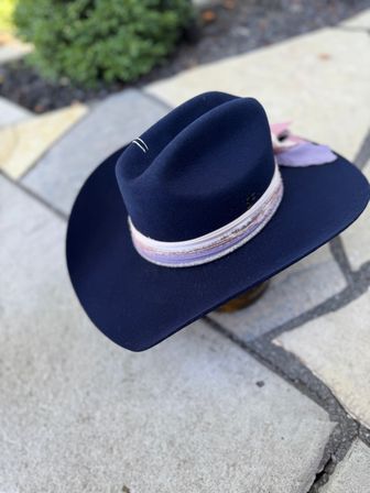 Navy felt wide‑brim cowboy hat with pastel pink and lavender ribbon and feather, resting on a sunlit stone patio