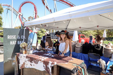 Smiling vendor at an outdoor hat stall under a white canopy, displaying stacked cowboy hats, cowhide-covered counter and accessories with a looping red roller coaster and sunny amusement-park greenery in the background.