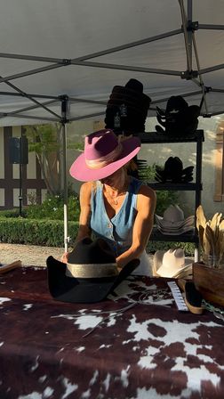 Sunlit outdoor market stall showcasing western hats — a seller in a purple fedora arranges black cowboy hats on a cowhide-covered table beneath a pop-up canopy.