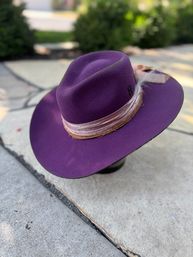 Purple wide-brim felt fedora with layered velvet ribbon and feather accent resting on a stone patio, stylish fashion accessory
