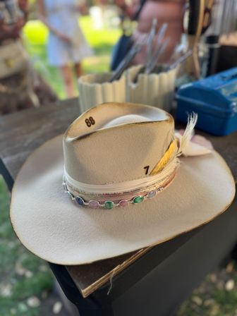 Sunlit beige felt cowboy hat with a feather and colorful gemstone band perched on a wooden table at an outdoor market