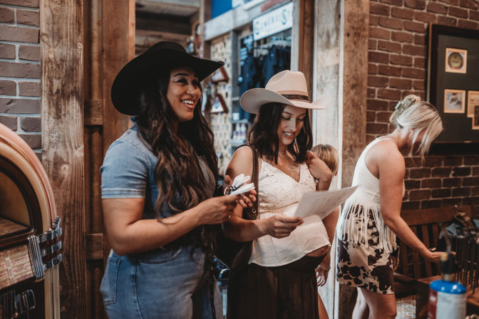 Two women in cowboy hats smiling and reading a paper while browsing a rustic western-wear shop with brick walls, wooden beams and a vintage jukebox