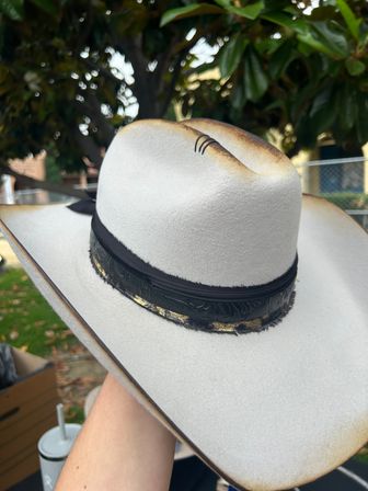 White felt cowboy hat with a wide brim and burnished brown edges, topped with a black tooled leather hatband with gold accents, held by a hand outdoors in a leafy backyard.