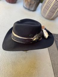 Black wide-brim felt western hat with stitched crown, decorative leather band, brass concho and feather, photographed on a neutral outdoor surface