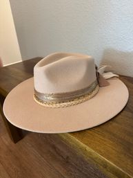 Beige wide-brim felt fedora with braided leather and gold chain band resting on a wooden table indoors