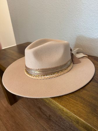 Beige wide-brim felt fedora with braided leather and gold chain band resting on a wooden table indoors