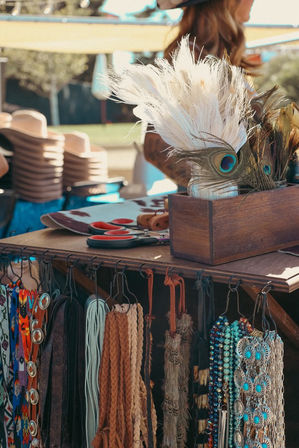 Sunlit outdoor boho market stall with peacock feathers in a wooden box, stacks of hats, scissors, hanging leather belts, braided straps and turquoise beaded necklaces.